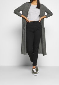 Grå ribbad cardigan med långa ärmar, grå kort topp, högmidjade svarta jeans med knappar och gröna sneakers. Enkla men funktionella kläder.