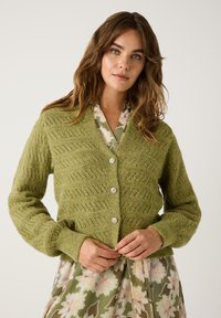Groene gebreide cardigan met een textuurpatroon, met een V-hals, lange mouwen en vijf decoratieve knopen. G gedragen over een bloemenjurk.
