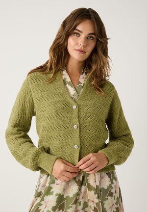 Cardigan en laine verte tricoté avec un motif texturé, avec un décolleté en V, des manches longues et cinq boutons décoratifs. Porté par-dessus une robe fleurie.