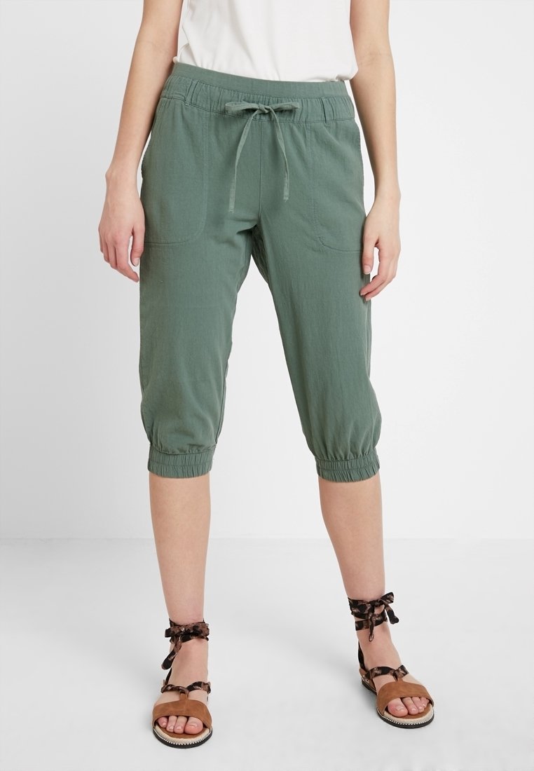Pantalones de cultivo verdes y ligeros con cintura elástica y lazo. Cuenta con dos bolsillos frontales y puños fruncidos. Textura suave.