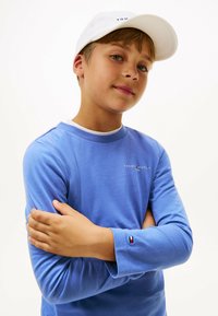 Blaues Langarmshirt mit Rundhalsausschnitt, das ein kleines Logo auf der Brust und einen kontrastierenden Ärmelabschluss in Rot, Weiß und Blau aufweist. Weiße Mütze.