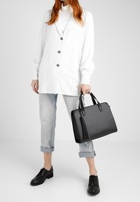 Borsa strutturata nera con finitura testurizzata, doppio manico e accentuazione con nappina. Indossata con un blazer bianco a righe sottili e jeans chiari risvoltati.