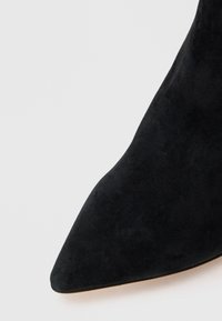 Chaussure noire en daim à bout pointu avec une texture lisse et une silhouette élégante. La semelle est d'une couleur beige clair, contrastant avec le matériau supérieur.