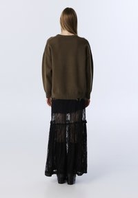 Brauner Strickpullover in lockerer Passform, kombiniert mit einem schwarzen Spitzen-Maxikleid, das ein gestuftes Design und einen durchsichtigen Überzug aufweist.