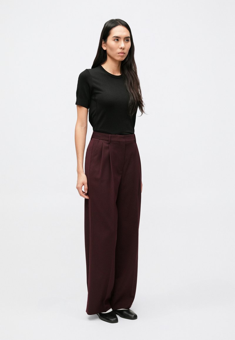 Black short-sleeve shirt paired with high-waisted, wide-leg burgundy trousers. Model stands on a plain background, wearing black shoes.
