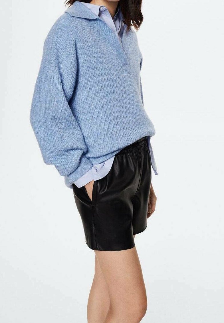 Femme portant un pull en maille bleu clair par-dessus une chemise à col, assorti à un short en cuir noir, debout les mains dans les poches.