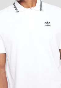 Man som bär en vit Adidas polo med svarta dubbelremsor på kragen och Adidas-logotypen på vänster bröst.