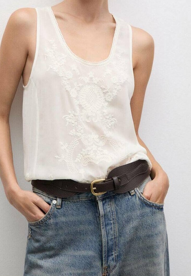 Bez rękawów, biała haftowana bluzka wsunięta w wysokiej talii niebieskie dżinsy, z ciemnobrązowym skórzanym paskiem i rękami w kieszeniach.