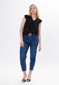 Sort ærmeløs top med flæser på skuldrene og tekstureret stof, parret med tætsiddende blå jeans og sorte åbne hæle; sølvbælte som accent.