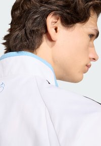 Νέος άνδρας με καστανά μαλλιά, φορόντας άσπρο μπουφάν με γαλάζια διακόσμηση, σε πλάγια όψη μπροστά σε απλό φωτεινό φόντο.