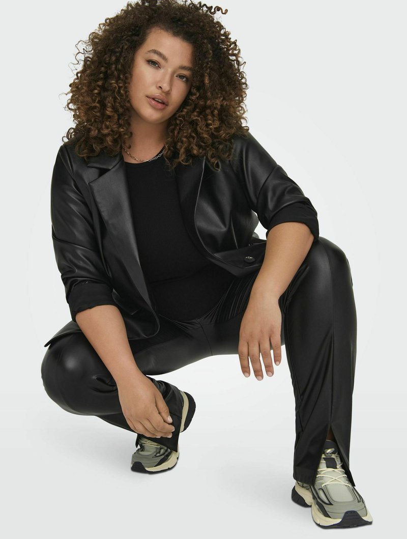 Vrouw met krullend haar in een zwart faux leren blazer en broek die hurkt, poseert met lichtgrijze en zwarte sneakers tegen een effen achtergrond.