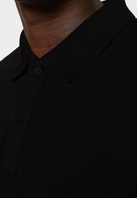 Polo shirt noir avec un design en tricot texturé, patte de boutonnage à deux boutons et col en ribbed. Mise au point sur la partie supérieure du vêtement.