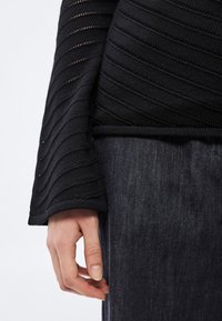 Fekete kötött felső texturált, ferde mintákkal és bővülő ujjakkal, sötét farmerrel párosítva. Látható kéz nyugszik a textilen.