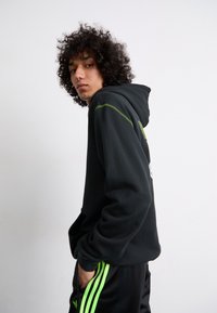 Svart hoodie med dragsko i huva, med gröna sömsmycken. Buren med svarta träningsbyxor som har vertikala gröna ränder.