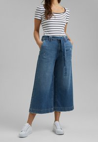 Vrouw met wijde, cropped blauwe spijkerbroek met een ceintuur, gestreept T-shirt met korte mouwen en witte sneakers, staand met handen in de zakken.