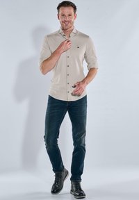 Mann in beiger, knopfgeknöpfter Bluse mit hochgekrempelten Ärmeln, dunklen Jeans und schwarzen Lederstiefeln, der vor einem schlichten, hellen Hintergrund steht.