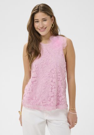 Młoda kobieta uśmiechająca się, ubrana w przezroczystą, różową, kwiecistą, koronkową bluzkę bez rękawów z dużym różowym kwiatem na ramieniu oraz białe spodnie.