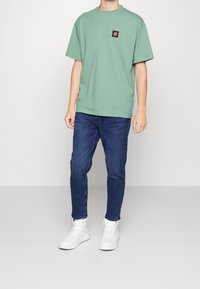 Zielona bawełniana koszulka z okrągłym dekoltem i naszywką na piersi, zestawiona z ciemnoniebieskimi dżinsami i białymi trampkami.