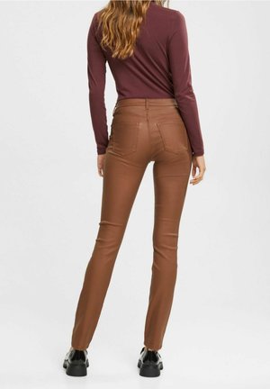 Pantaloni marroni aderenti con una texture liscia e cinque tasche, abbinati a un top a maniche lunghe color marrone scuro. Le calzature presentano una punta rotonda e una suola spessa.