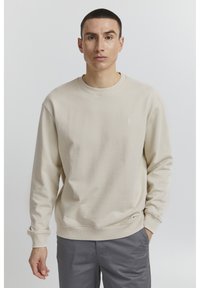 Sudadera beige con cuello redondo, puños y dobladillo de canalé. Presenta un pequeño detalle de logo en el pecho. Combinada con pantalones grises.