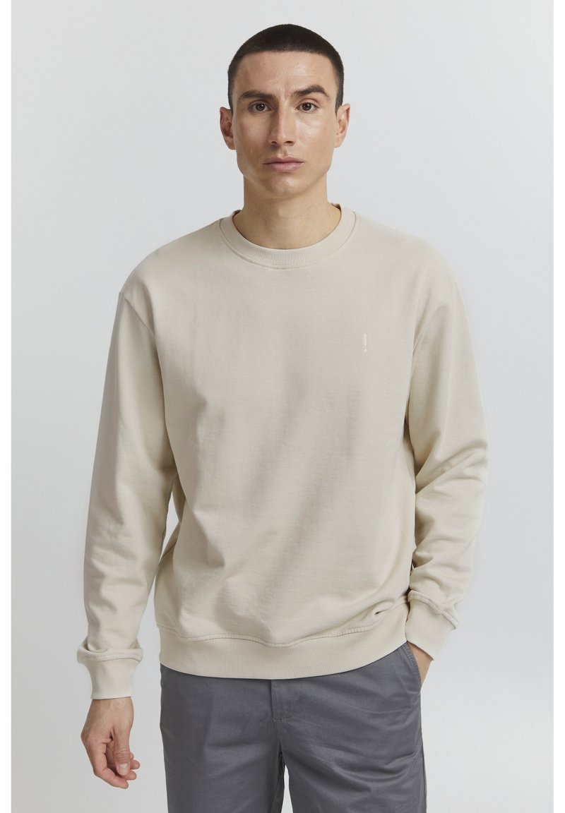 Sudadera beige con cuello redondo, puños y dobladillo de canalé. Presenta un pequeño detalle de logo en el pecho. Combinada con pantalones grises.