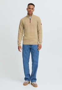 Beige gebreid sweater met een halve ritssluiting, geribde manchetten en zoom. Gepaard met blauwe denim jeans en beige sneakers met lichte accenten.