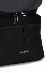 Sac à dos noir en tissu durable, avec un rabat supérieur, un compartiment avant zippé et un logo Volcom bien en vue sur le bas de l'avant.
