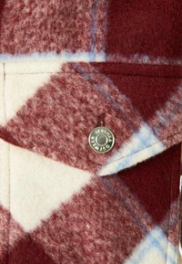 Wzór w kratę z wełny, z białymi i burgundowymi kwadratami oraz niebieskimi akcentami. Widziany z bliska srebrny zatrzask znajduje się blisko kieszeni.