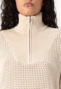 Beige, ribbet strikket genser med høy krage og halvmeter glidelås. Har et mesh-mønster på ermene og brystområdet.