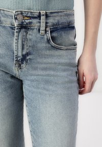 Vaaleansiniset farkut, joissa on kapea leikkaus, näkyvät saumat, viisi taskua ja nappikiinnitys vyötäröllä.