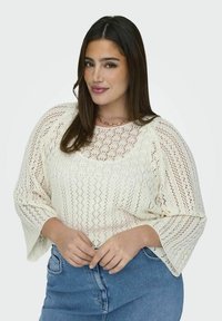 Strick-Oberteil in Creme mit Spitzenmuster, weiten Ärmeln und rundem Halsauschnitt, kombiniert mit hochgeschnittenen Jeans aus Denim.