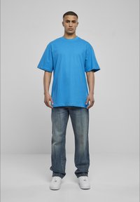 Cămașă din bumbac de culoare albastru deschis, cu un croi lejer și mâneci scurte, asortată cu blugi din denim decolorat și adidași albi. Se află în fața unui fundal simplu.