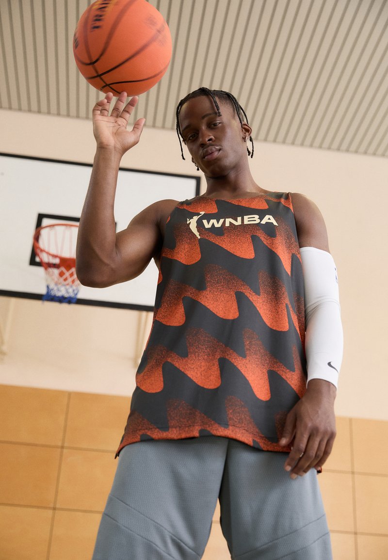 Camiseta de tirantes de la WNBA en naranja y negro, con un patrón de olas. Combinada con pantalones cortos grises y una manga blanca, sujetando un balón de baloncesto por encima.