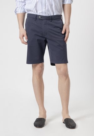 Hombre con pantalones cortos azul marino hasta la rodilla, una camisa blanca y azul a rayas con las mangas remangadas y zapatos negros tejidos sin cordones.