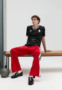 Chemise noire à manches courtes avec une texture brillante et un motif blanc, associée à un pantalon rouge ample avec des rayures latérales. Chaussures noires avec détail de sangle.