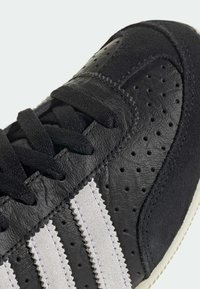 Black sneaker with perforated texture, suede accents, and gray stripes. Features leather material and black laces. White sole.