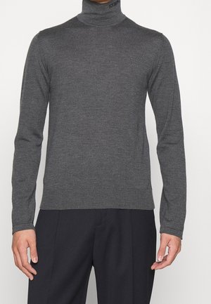 Mann trägt einen eng anliegenden dunkelgrauen Rollkragenpullover mit dezentem Markenschriftzug am Kragen, kombiniert mit schwarzer Hose vor einfarbigem Hintergrund.