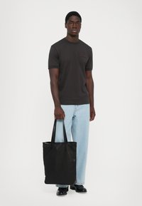 Mann står med en svart t-skjorte, lyseblå denim, svarte sko, holder en stor svart totebag mot en enkel hvit bakgrunn.
