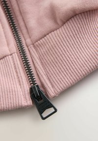 Světle růžový zip-up oděv s vroubkovanou texturou na lemu, s kontrastním černým zipem a detaily z kovu.