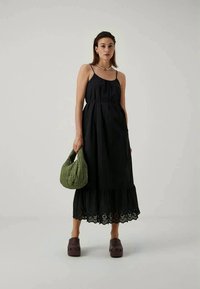 Robe sans manches noire avec un ourlet en couches et des détails en œillets, associée à un sac matelassé vert et des sabots marron.