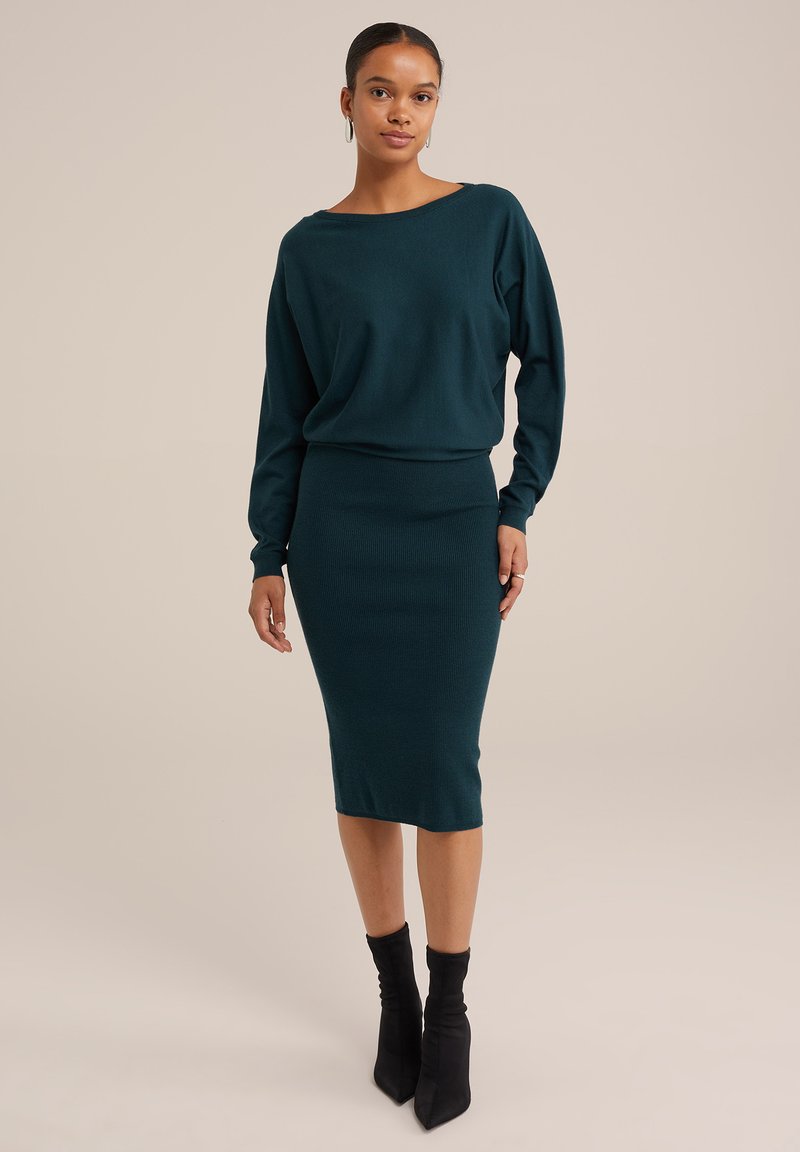 Turkizni pletenini pulover z ladijskim izreza in dolgimi rokavi, v kombinaciji z ujemajočo se prilegajočo midi krilom. Dodatki vključujejo srebrne obročaste uhane.