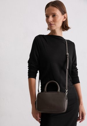 Woman wearing long-sleeve black dress with dark gray rectangular crossbody bag with top handles and adjustable strap.