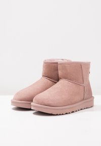 Un par de botas de invierno cortas de ante rosa claro con forro suave y suelas de goma estriadas sobre una superficie blanca.