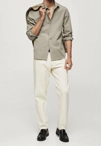 Chemise à manches longues boutonnée en tissu gris clair, portée avec un pantalon crème et des mocassins noirs. La chemise a les manches retroussées.