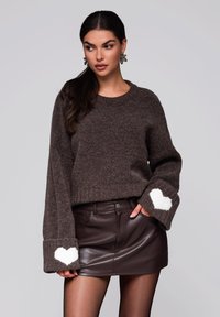 Brązowy sweter dzianinowy o prążkowanej strukturze z białymi akcentami serc na podwiniętych mankietach, zestawiony z brązową mini spódnicą z ekoskóry.