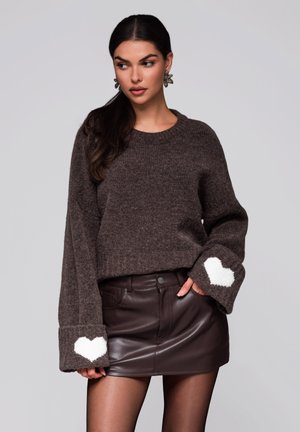 Braun gestrickter Pullover mit rippenförmiger Textur und weißen Herz-Akzenten an den umgeschlagenen Ärmeln, kombiniert mit einem braunen Fake-Leder Minirock.