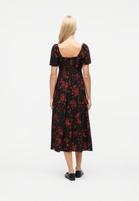Femme aux longs cheveux blonds portant une robe midi noire à imprimé floral rouge, col carré, manches bouffantes et chaussures plates noires, vue de dos.