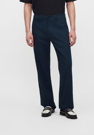 Homme portant un pantalon bleu marine droit et des mocassins noir et blanc avec un haut noir sur un fond uni.