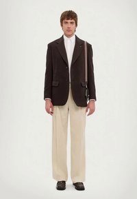 Homem a usar um blazer castanho escuro, camisa branca, calças largas beiges, sapatos pretos e uma pequena mala de ombro preta com uma alça riscada.