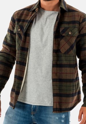 Mężczyzna ubrany w rozpiętą brązowo-zielono-czarną kraciastą kurtkę na jasnoszarej koszulce i niebieskich dżinsach.